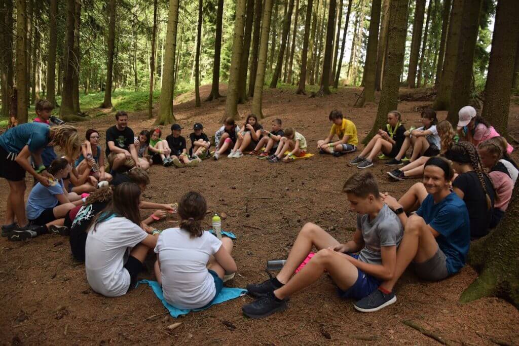 Na obrázku jsou děti s lektorem sedící v kruhu v lese.